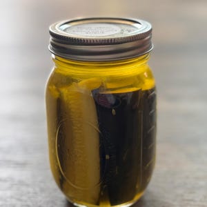 Peut inclure: Un bocal en verre rempli de saumure jaune et de deux cornichons entiers à l'aneth. Le bocal a un couvercle argenté avec une étiquette blanche.