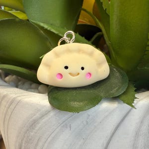 May include: A white clay charm shaped like a dumpling with a smiling face and pink cheeks. The charm is hanging from a silver loop.