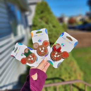 May include: Three PopSockets phone grips featuring a gingerbread Mickey Mouse design. The grips are packaged in clear plastic with the PopSockets logo. The gingerbread Mickey Mouse designs are decorated with red and green icing and sprinkles.