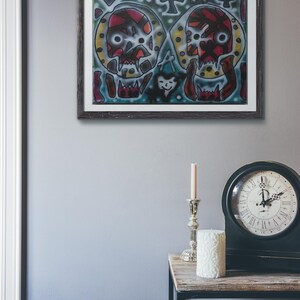 May include: A framed abstract painting with two colorful skulls in the center. The background is a blue and green color with various shapes and symbols.