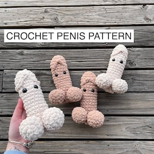 May include: Four handmade crochet penis toys in varying shades of beige and tan, each with black button eyes. The text "CROCHET PENIS PATTERN" is displayed in a white rectangle above the toys. The toys are arranged on a wooden surface.