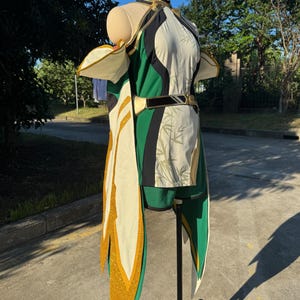 Puede incluir: Un disfraz sin mangas que presenta un corpiño color crema con un sutil patrón de hojas de bambú, acentuado por paneles laterales de color verde oscuro y un cuello alto dorado. El conjunto incluye pantalones cortos verdes a juego, un cinturón dorado y paneles de capa largos de color crema con ribete de purpurina dorada.