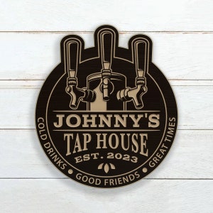 May include: A black and brown round wooden sign with a vintage style illustration of three beer taps. The sign reads "Johnny's Tap House Est. 2023 Cold Drinks Good Friends Great Times".