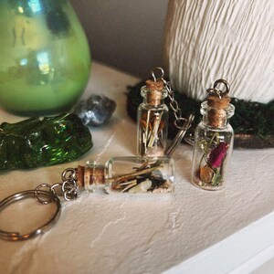 May include: Three small glass bottles with cork tops and silver chains attached. Each bottle is filled with dried flowers, herbs, and other natural elements. The bottles are arranged on a white surface.