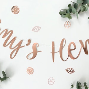 May include: Rose gold banner with the text "Amy's Hen" in a cursive font. The banner is decorated with small rose gold flowers and leaves.