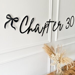 May include: Black banner with the words "Chapter 30" in a cursive font, with a bow on the left side. The banner is hanging on a white wall. A clear glass vase with dried flowers sits on a wooden table.