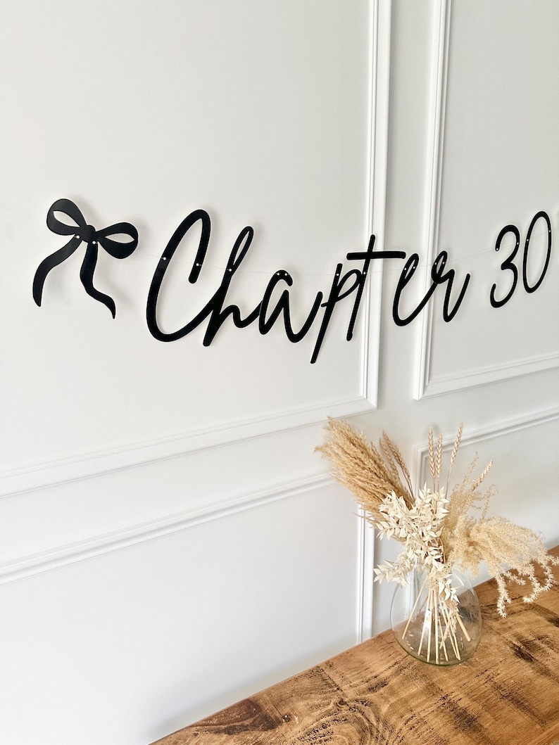 May include: Black banner with the words "Chapter 30" in a cursive font, with a bow on the left side. The banner is hanging on a white wall. A clear glass vase with dried flowers sits on a wooden table.