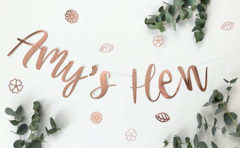 May include: Rose gold banner with the text "Amy's Hen" in a cursive font. The banner is decorated with small rose gold flowers and leaves.