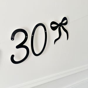 May include: Black metal number 30 with a bow accent, perfect for a birthday or anniversary celebration. The numbers are in a bold, cursive font, and the bow adds a decorative touch. Ideal for party decorations.