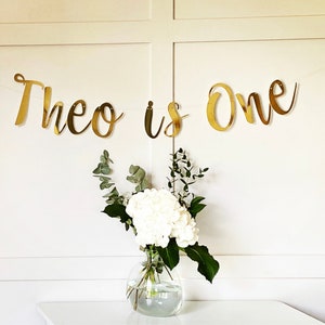May include: Gold glitter banner with the text "Theo is One" hanging on a white wall. A vase with white flowers and green leaves sits on a white table in front of the banner.