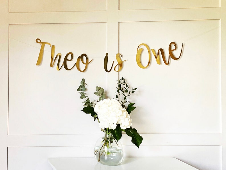 May include: Gold glitter banner with the text "Theo is One" hanging on a white wall. A vase with white flowers and green leaves sits on a white table in front of the banner.