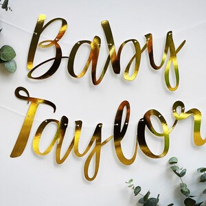 May include: Gold foil banner with the text "Baby Taylor" in a cursive font.