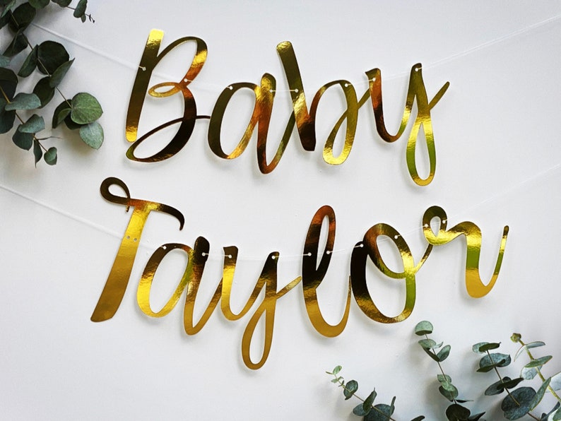 May include: Gold foil banner with the text "Baby Taylor" in a cursive font.