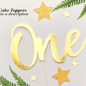 May include: Gold glitter cake topper with the word "One" in a cursive font. The topper is surrounded by small gold glitter stars.