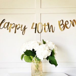 May include: Gold foil banner with the text "Happy 40th Ben" on a white wall with white flowers in a vase.