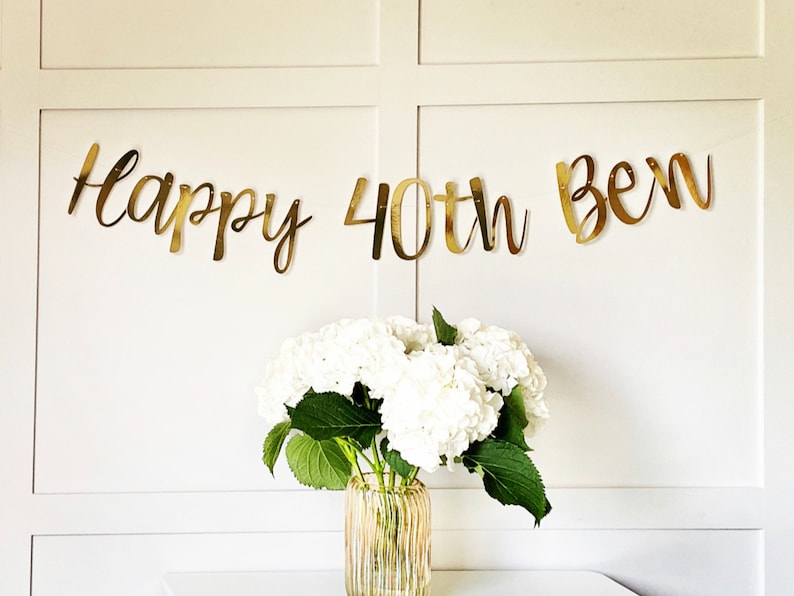 May include: Gold foil banner with the text "Happy 40th Ben" on a white wall with white flowers in a vase.