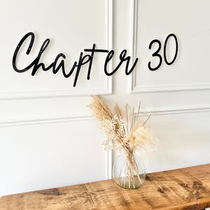 May include: Black cursive "Chapter 30" banner displayed on a white wall. A clear glass vase with dried flowers sits on a wooden table. The banner is a decorative element for a milestone birthday.