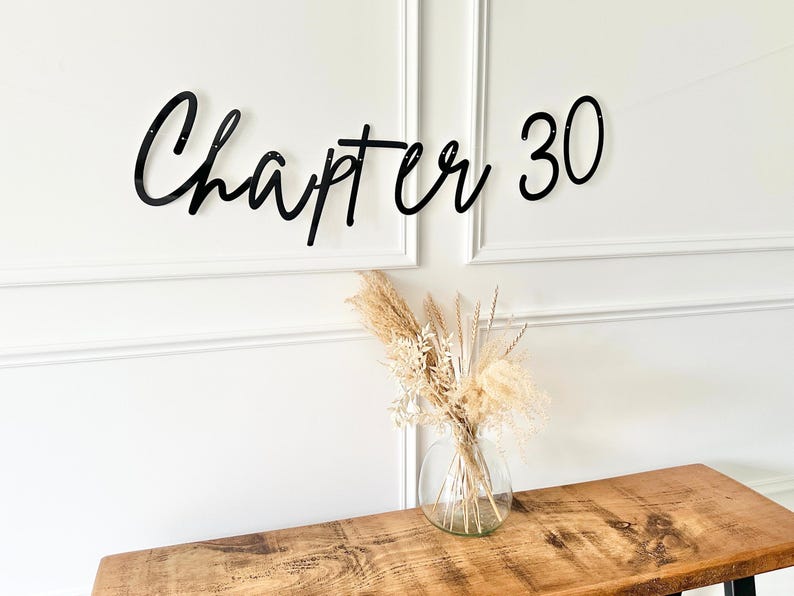 May include: Black cursive "Chapter 30" banner displayed on a white wall. A clear glass vase with dried flowers sits on a wooden table. The banner is a decorative element for a milestone birthday.