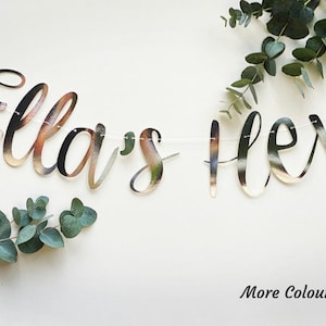 May include: A silver glitter banner with the text "Ella's Hen" in cursive script. The banner is strung with white thread and is decorated with eucalyptus leaves.