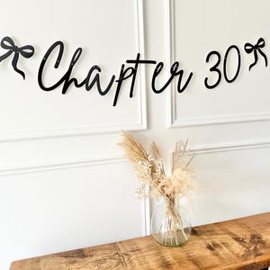 May include: Black cursive banner that reads "Chapter 30" with bow accents. The banner is hanging above a wooden table with a clear glass vase filled with dried flowers. The wall is white with decorative molding.