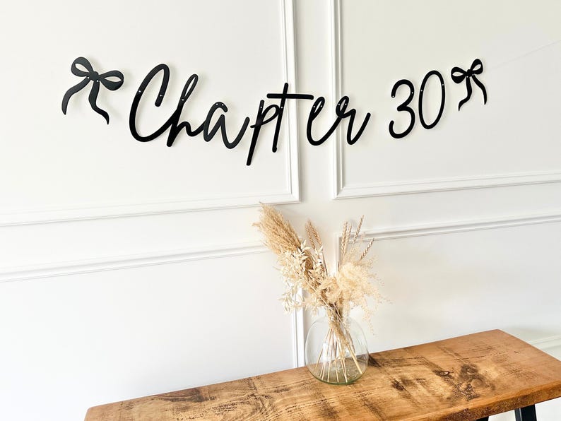 May include: Black cursive banner that reads "Chapter 30" with bow accents. The banner is hanging above a wooden table with a clear glass vase filled with dried flowers. The wall is white with decorative molding.