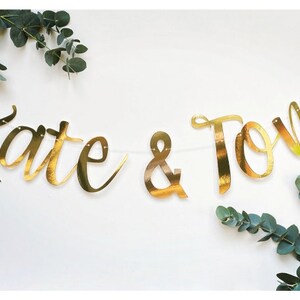 May include: Gold foil banner with the text "Kate & Tom" hanging between two sprigs of eucalyptus.