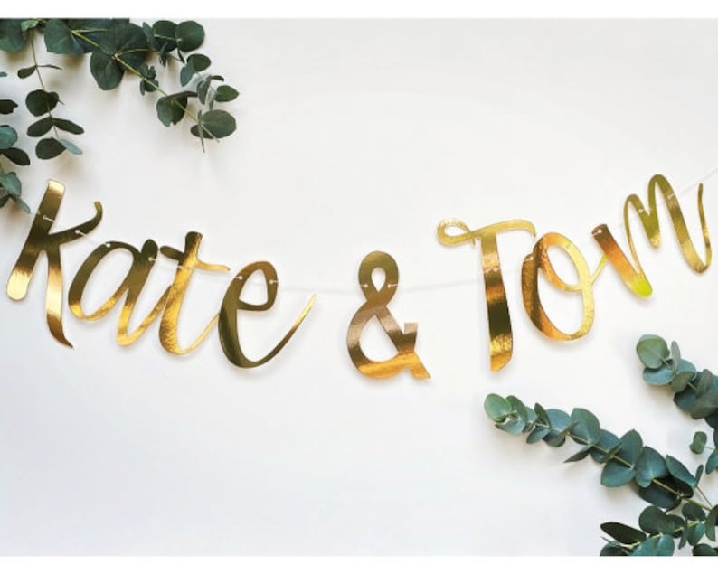 May include: Gold foil banner with the text "Kate & Tom" hanging between two sprigs of eucalyptus.