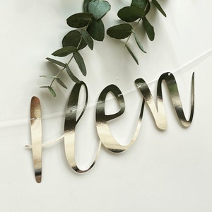 May include: A silver foil banner with the word "flen" written in cursive script. The banner is hanging from a clear string and is decorated with eucalyptus leaves.