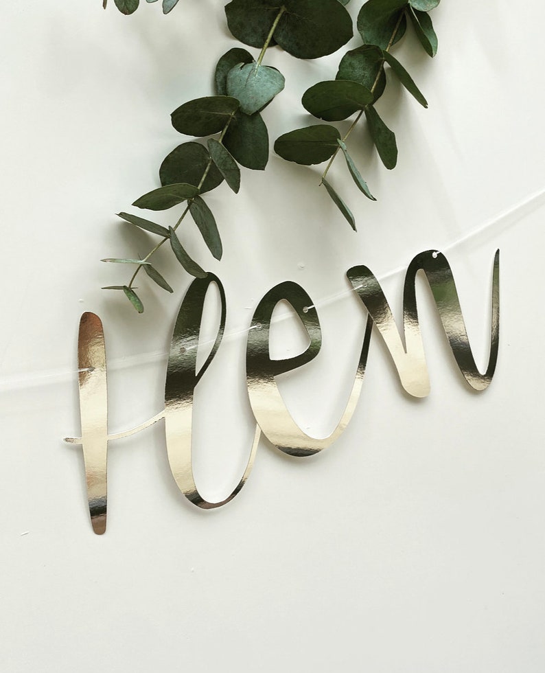 May include: A silver foil banner with the word "flen" written in cursive script. The banner is hanging from a clear string and is decorated with eucalyptus leaves.