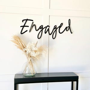 May include: Black metal sign with the word "Engaged" in a cursive font. The sign is hanging on a white wall above a black console table with a vase of dried flowers.