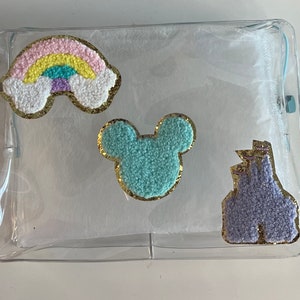 Op de afbeelding: Een doorzichtige plastic tas met rits en drie vilt applicaties: een regenboog, een Mickey Mouse hoofd en een paars kasteel. De applicaties zijn omrand met gouden glitter.