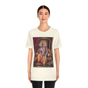May include: A white t-shirt with a colorful image of a Hindu deity sitting on a throne. The deity is wearing a blue robe, a yellow sash, and a crown. The throne is decorated with gold and red.