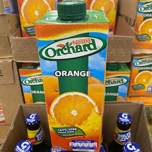May include: A carton of Nestle Orchard orange juice with a green lid. The carton is sitting on a shelf with other cartons of orange juice and a six-pack of Malta Goya.