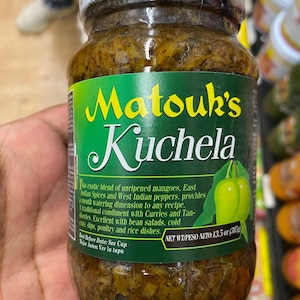 May include: A jar of Matouk's Kuchela, a traditional condiment. The label features green and yellow text with a graphic of green mangoes. The jar contains a blend of mangoes, spices, and peppers. Net weight is 13.5 oz.