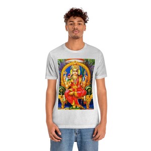 May include: A white t-shirt with a colorful image of a Hindu goddess sitting on a throne. The goddess is wearing a red and gold sari and has four arms. The background is a blue and gold design.