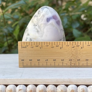 May include: A polished, egg-shaped stone with a white and lavender pattern. A wooden ruler shows the stone is approximately 3 inches tall. The stone sits on a white surface, with a blurred green background.