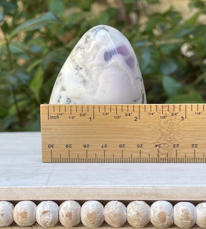 May include: A polished, egg-shaped stone with a white and lavender pattern. A wooden ruler shows the stone is approximately 3 inches tall. The stone sits on a white surface, with a blurred green background.