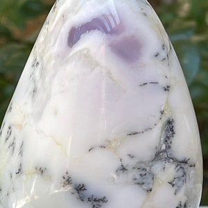 May include: A polished, egg-shaped stone with a white base and intricate patterns. The stone features swirling white and gray veining, with delicate black dendrites resembling tiny ferns. A soft lavender hue is visible near the top, adding a touch of color.