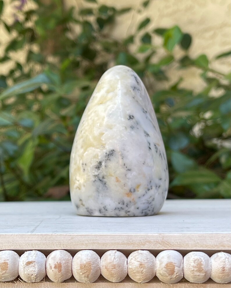 May include: A smooth, teardrop-shaped stone with a cream-colored base and speckled black and gray patterns. The polished surface reflects light, highlighting the natural variations in the stone. The stone is displayed on a white wooden surface.