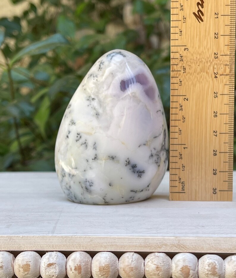 May include: A polished, egg-shaped stone with a white base, featuring dendritic patterns in black and hints of purple. A wooden ruler is placed next to the stone, showing measurements in inches.