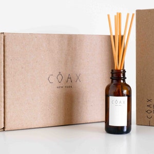 May include: A brown cardboard box with the text "COAX NEW YORK" printed on it. A brown glass bottle with a white label that says "COAX NEW YORK" is in front of the box. The bottle has a set of wooden sticks in it.