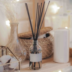 Könnte beinhalten: Eine klare Glasflasche mit schwarzen Diffusorstäbchen und einem weißen Etikett, auf dem "COAX" in schwarzer Schrift steht. Die Flasche steht auf einer Holzoberfläche mit einem Weidenkorb und einer weißen Kerze.