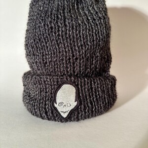 Könnte beinhalten: Eine dunkelgraue Strickmütze mit umgeschlagenem Saum. Die Mütze hat einen weißen Aufnäher mit einem stilisierten Alien-Kopf-Design. Die Mütze ist aus dickem, strukturiertem Garn gefertigt, das für kaltes Wetter geeignet ist.