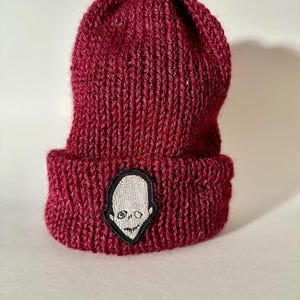 Könnte beinhalten: Eine bordeauxrote Strickmütze mit einem weißen Totenkopf-Patch.