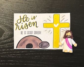 May include: A white card with the text "He is risen" and "He is risen indeed!" is next to a yellow cross with a heart and a small figurine of Jesus with a pink sash.