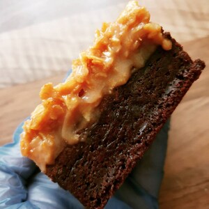 May include: A close-up of a brownie with coconut pecan frosting. The brownie is dark brown and the frosting is a light brown colour. The dessert is cut into a square shape.