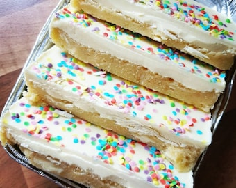May include: Four rectangular dessert bars with a shortbread base, a thick layer of white frosting, and colorful sprinkles. The bars are arranged in a silver foil container, showcasing a sweet treat.