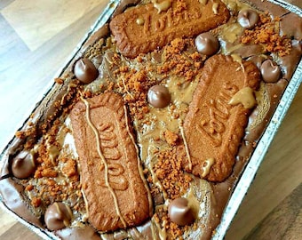 May include: A chocolate brownie topped with crumbled cookie crumbs, chocolate chips, and several whole cookies. The brownie is in a silver foil baking pan.