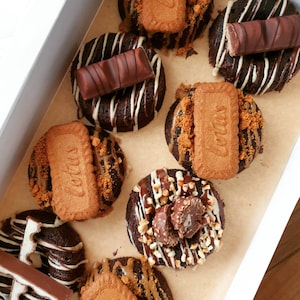 May include: A selection of chocolate donuts in a box. The dark brown donuts are decorated with white icing stripes and various toppings, including chocolate bars and biscuits. Some have crushed biscuit toppings.