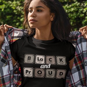 May include: Black t-shirt with the words "Black and Proud" spelled out using periodic table elements. The shirt is black with white text. The person in the image is wearing a plaid shirt over the t-shirt and blue jeans.